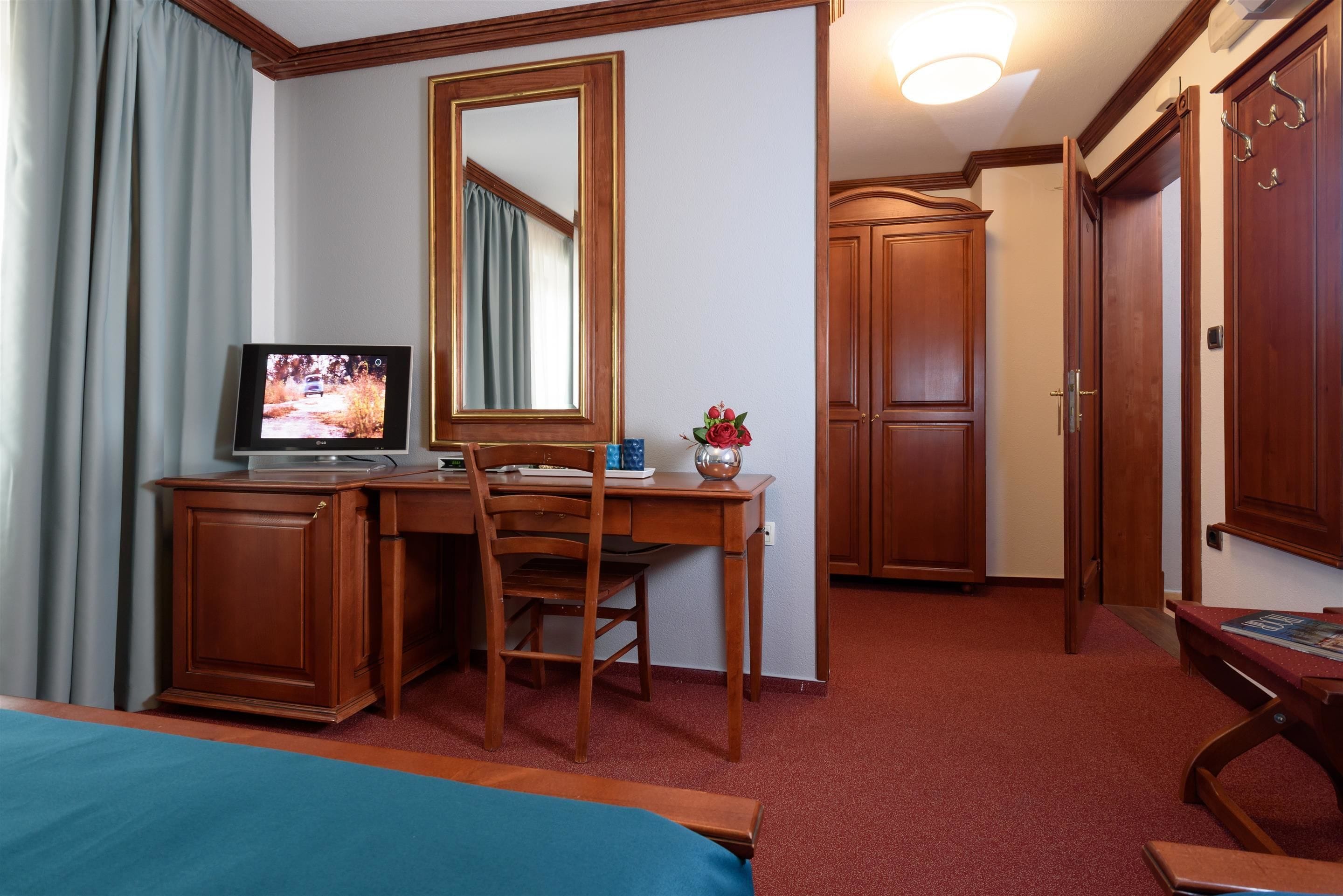 Une chambre d’hôtel avec un bureau en bois, une télévision, une chaise, un lit, un miroir et une porte.