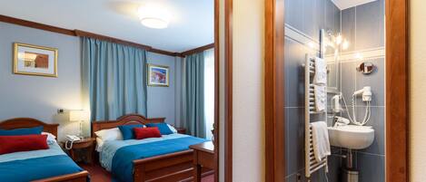 Une chambre d’hôtel avec deux lits en bois, une salle de bain avec un lavabo et un miroir, et un sol à carreaux.