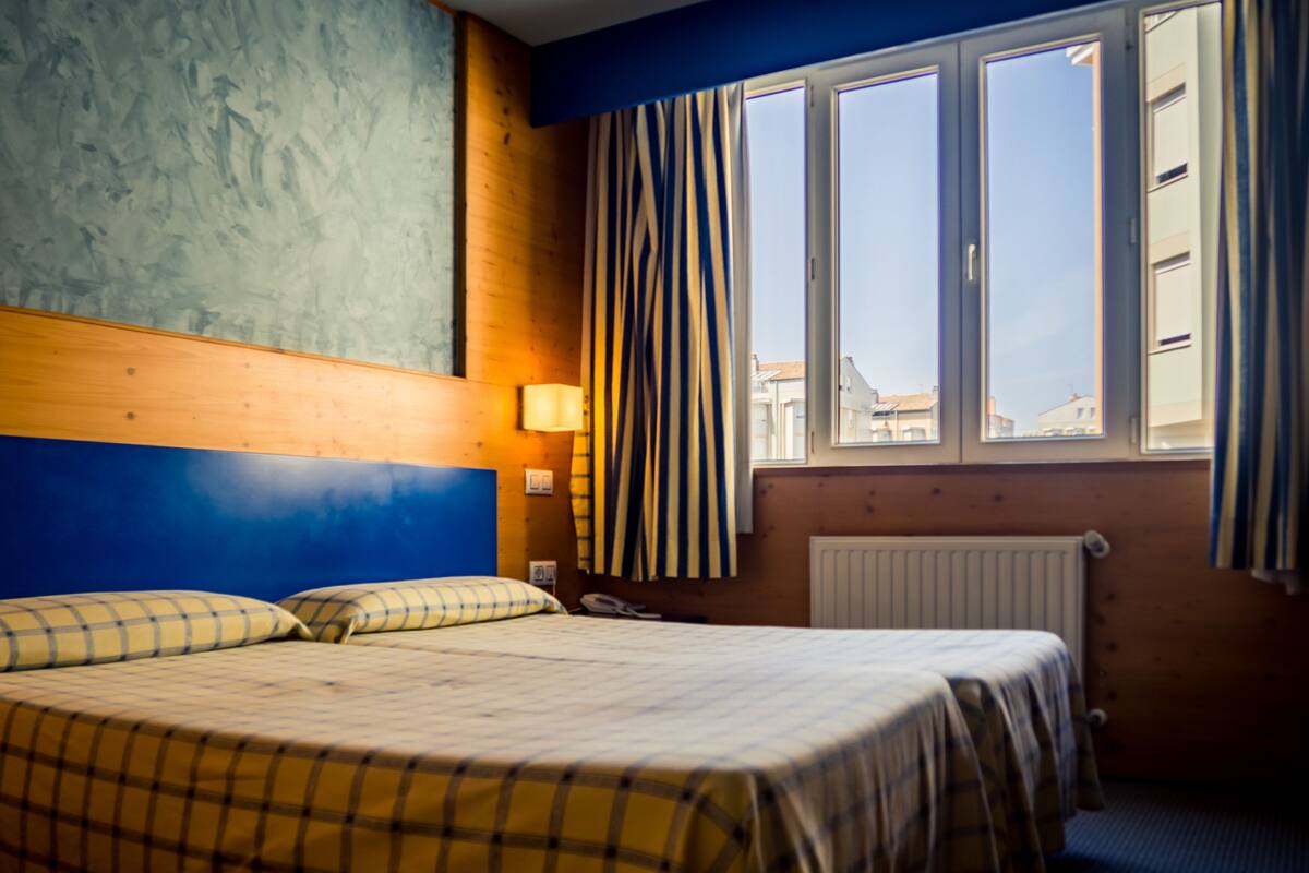 A hotel room with a bed, a window with curtains, a wall-mounted lamp, and a radiator.