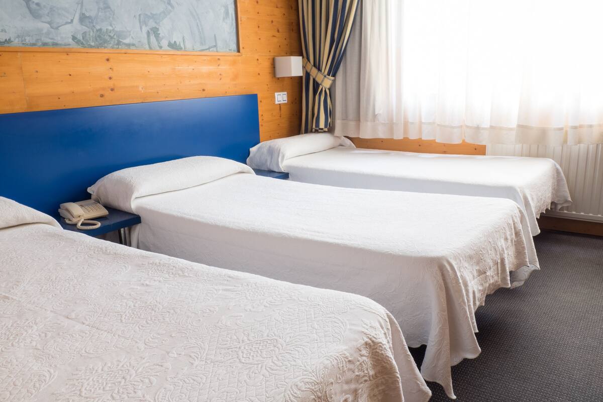 A hotel room with three single beds, a wooden headboard, a telephone on the nightstand, and a window with curtains.