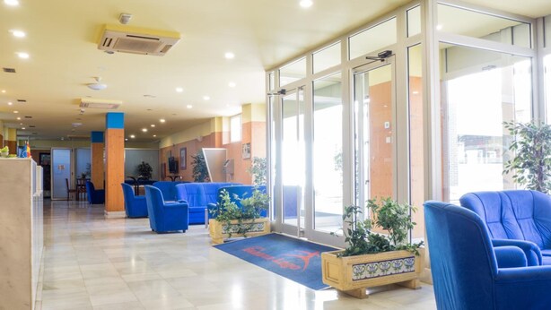 Un vestíbulo de hotel con mostrador de recepción, zona de estar azul y plantas en macetas.