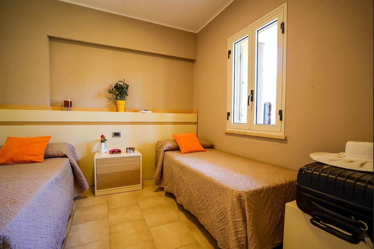 A small, cozy room with two single beds, a bedside table with a lamp, a potted plant, and a window with blinds.