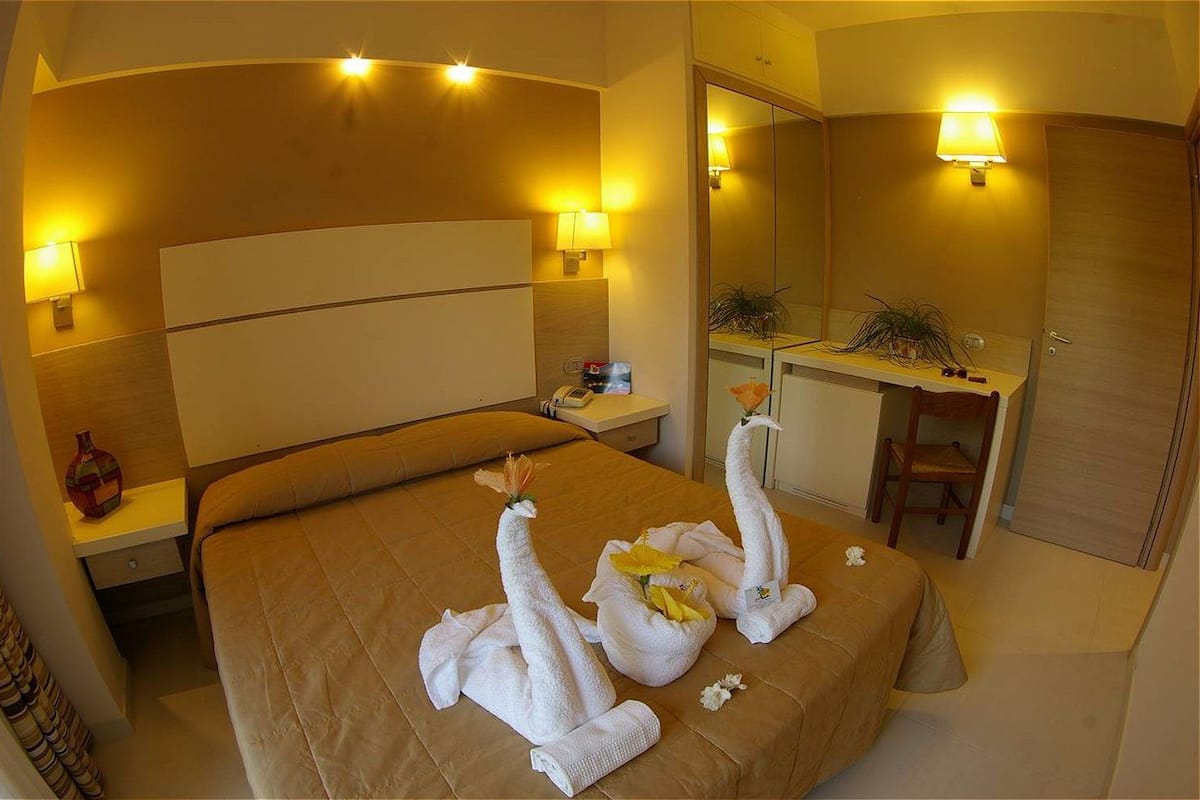 A hotel room with a large bed, two wall-mounted lamps, a bedside table, a mirror, and a wooden door.