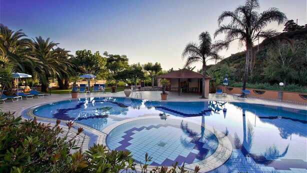 A pool area with lounge chairs and umbrellas, surrounded by palm trees and a small building. - Old River Residence (Ricadi)