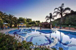 Un área de piscina con sillas de descanso y sombrillas, rodeada de palmeras y un pequeño edificio.