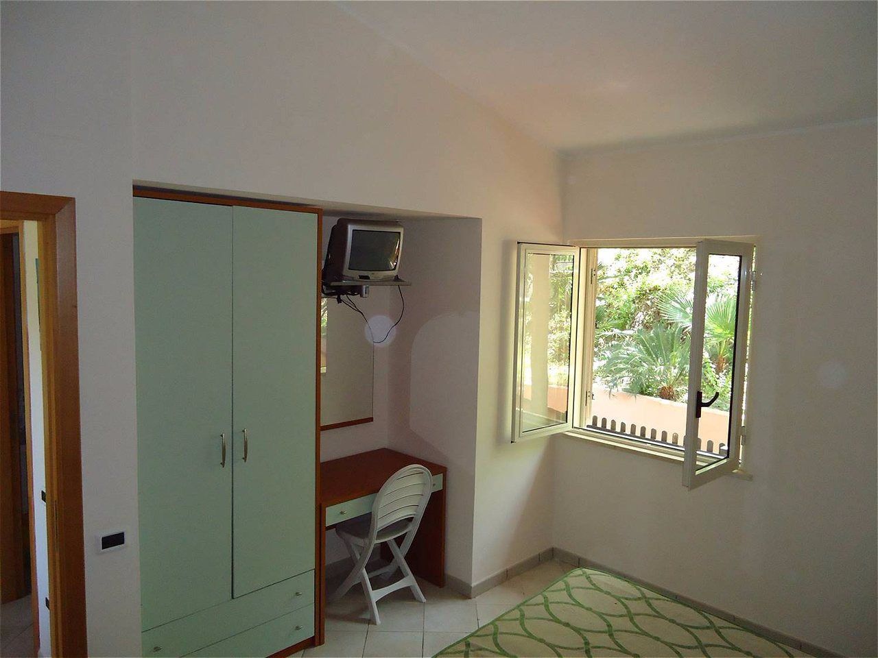 A room with a desk, chair, wardrobe, TV, and a window with a view of greenery.