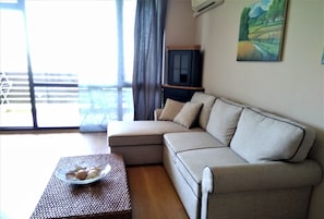 A living room with a beige sectional sofa, a small table with a bowl of fruit, a patterned ottoman, and a painting on the wall.