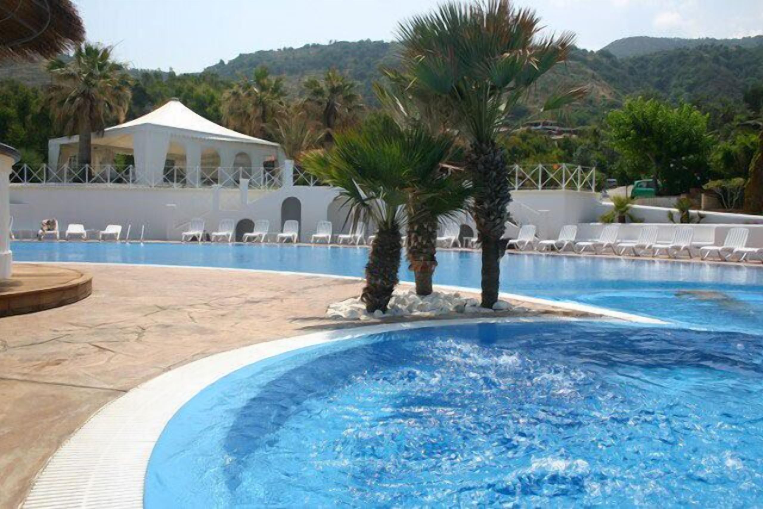 Piscina con sillas de descanso y palmeras, rodeada por un edificio blanco con arcos.