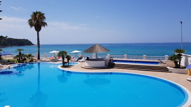 Piscina azul y cristalina con vistas al mar, rodeada de palmeras y un quiosco con techo de paja.