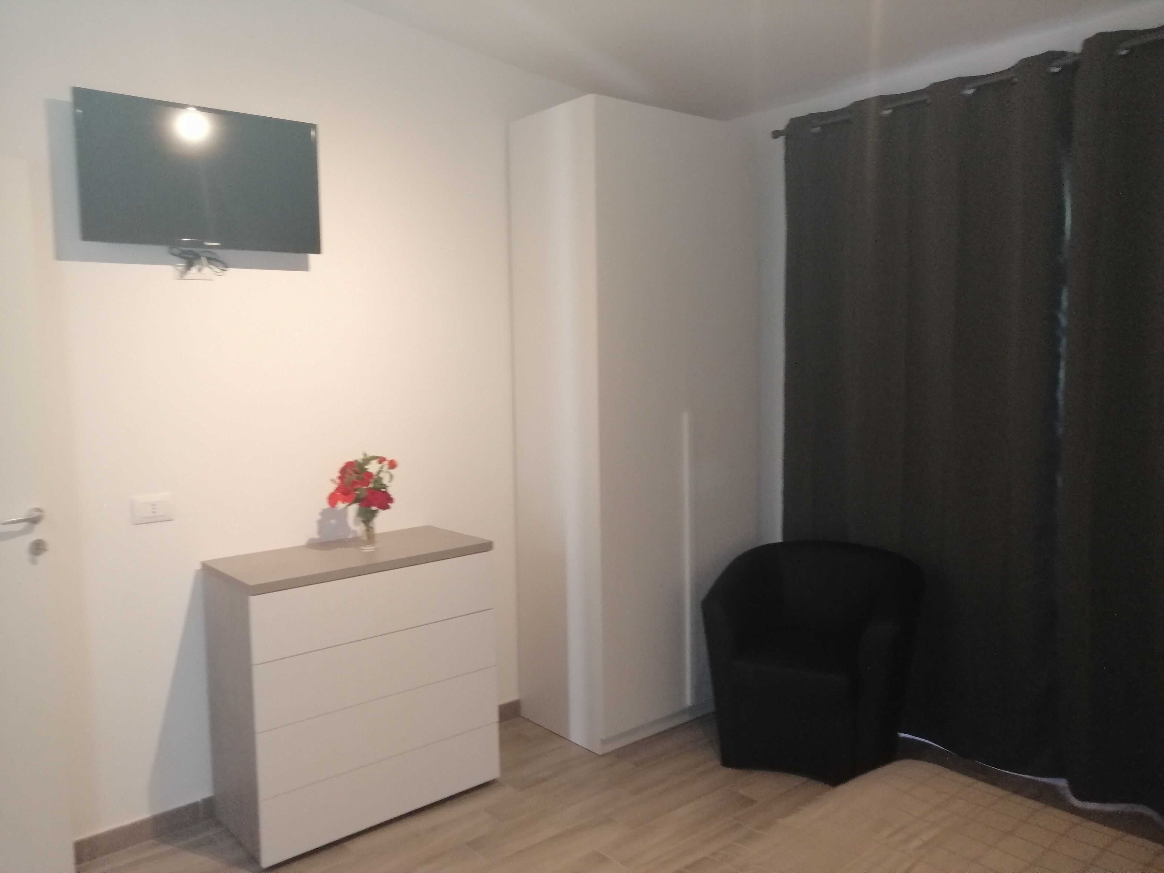 A small, modern room with a flat-screen TV mounted on the wall, a small white dresser, a black chair, and a dark curtain covering a window.