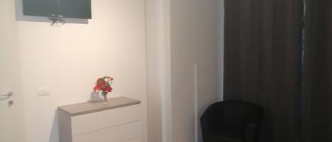 A small, modern room with a flat-screen TV mounted on the wall, a small white dresser, a black chair, and a dark curtain covering a window.