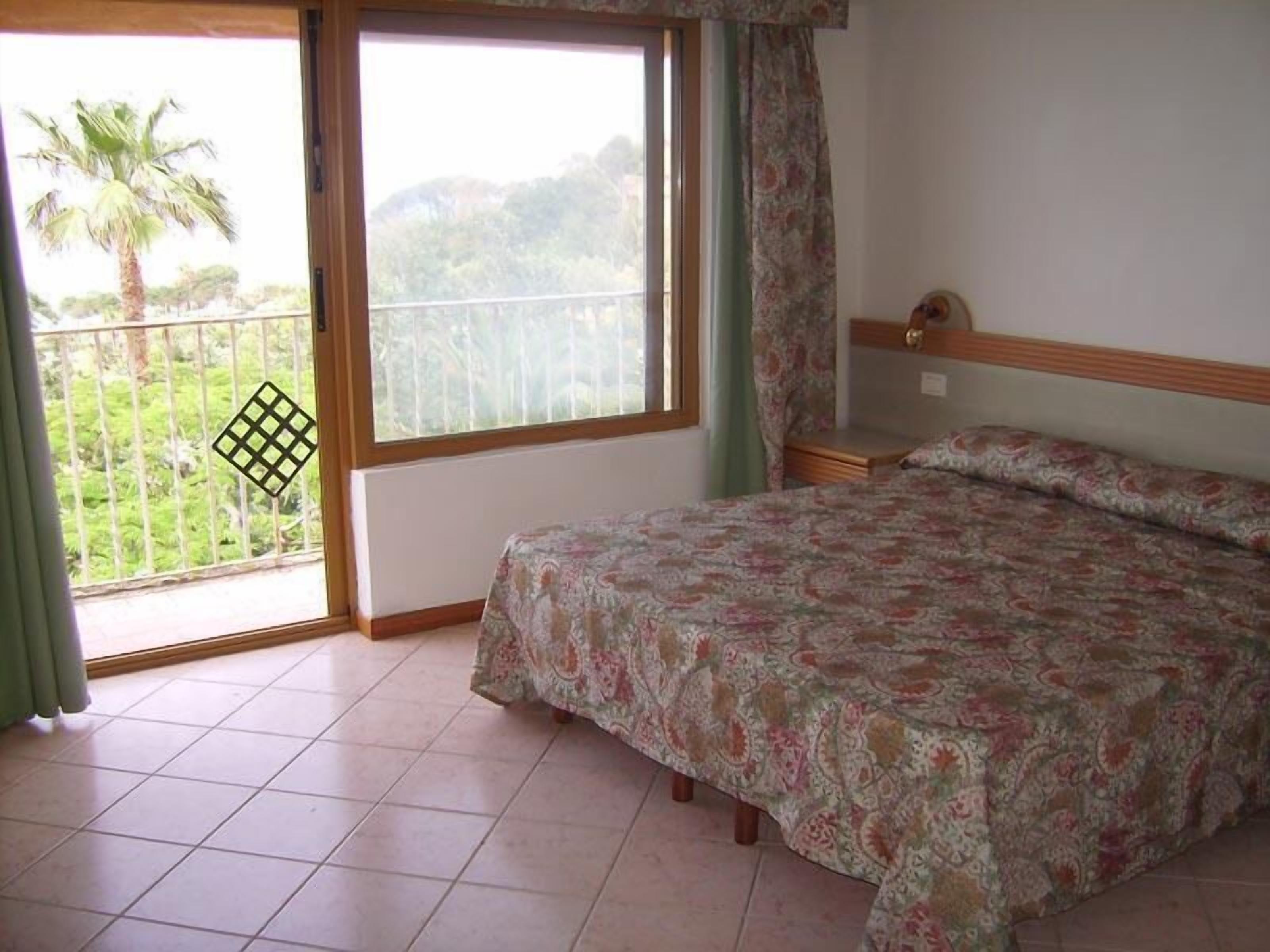 A bedroom with a bed, a window with a view of greenery, and a balcony with a railing.