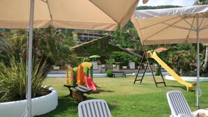 Zona de juegos para niños con toboganes y columpios, rodeada de sillones blancos y sombrillas.