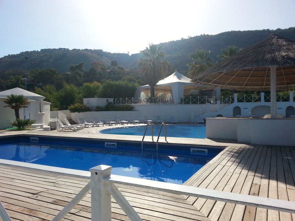Ein Poolbereich mit Liegestühlen, einem mit Stroh gedeckten Pavillon und Blick auf die Hügel im Hintergrund.