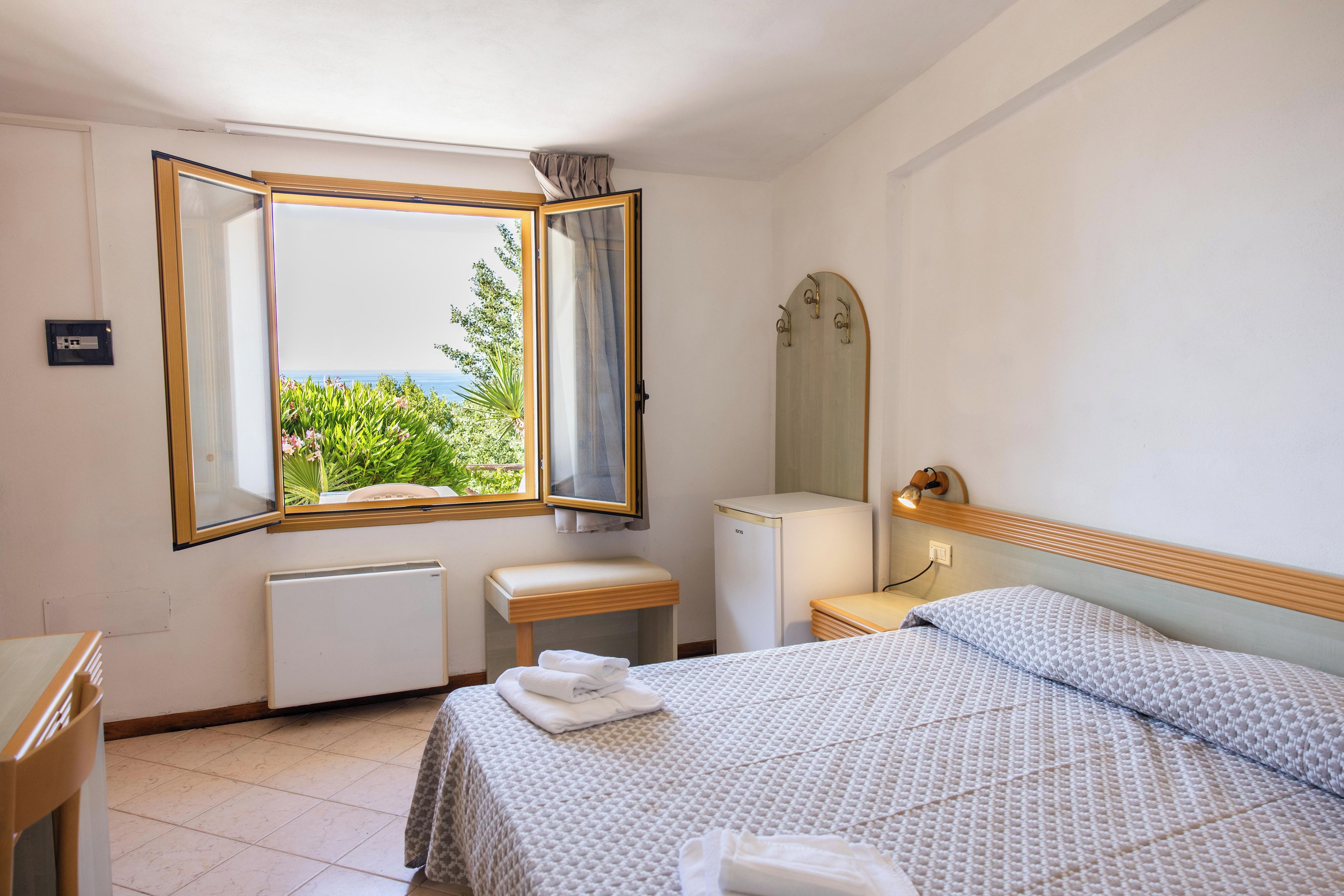A hotel room with a bed, a bedside table, a mirror, a radiator, and a window with a view of greenery and the sea.