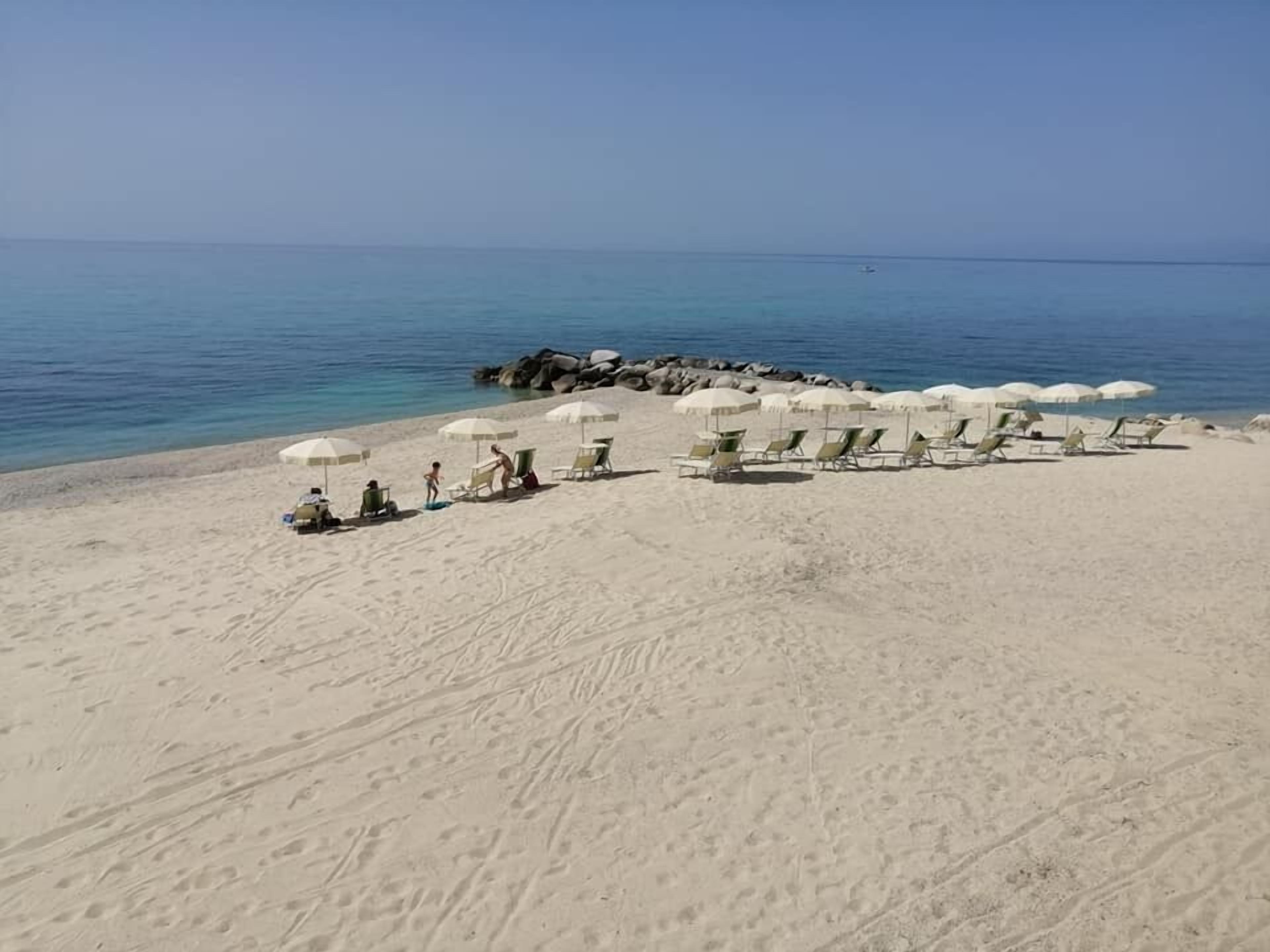Una playa con filas de tumbonas y sombrillas, costa rocosa y mar tranquilo.
