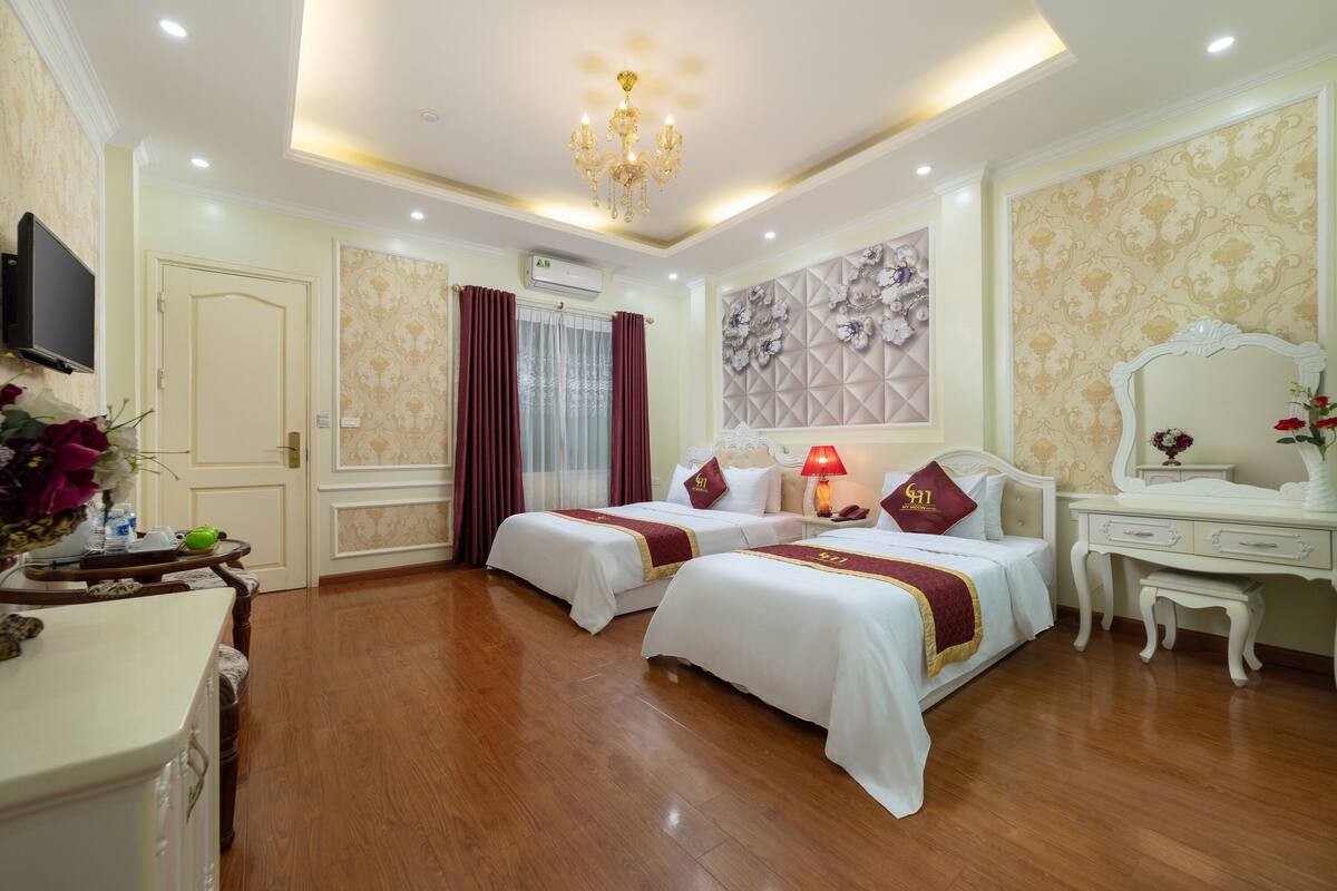 A hotel room with two beds, a desk, a chair, a TV, and a chandelier.