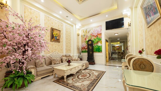 Un salon bien éclairé, avec un canapé, une table basse et une table à manger entourée de chaises. On y trouve également une grande plante en pot, ornée de fleurs roses, et une horloge fixée au mur.