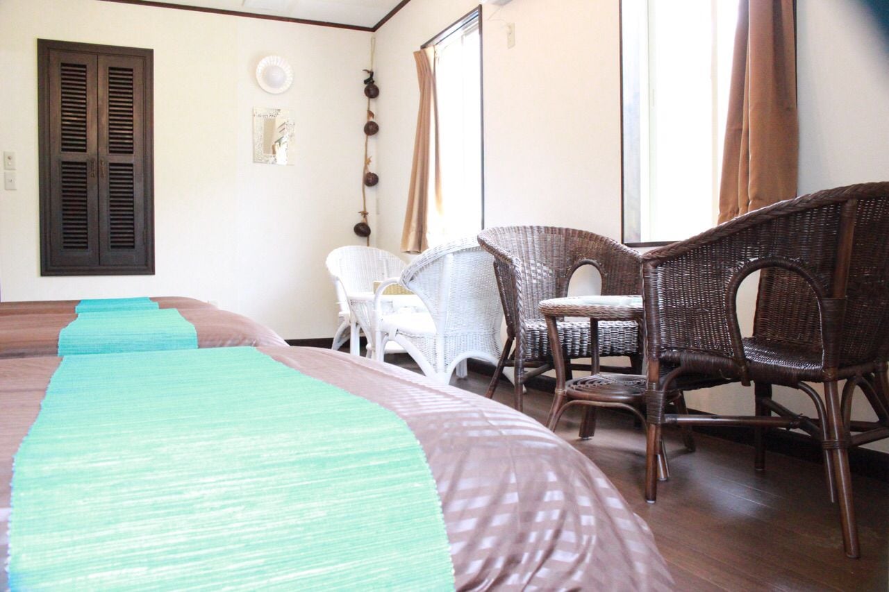 A hotel room with a large bed, two wicker chairs, a small table, and a window with curtains.