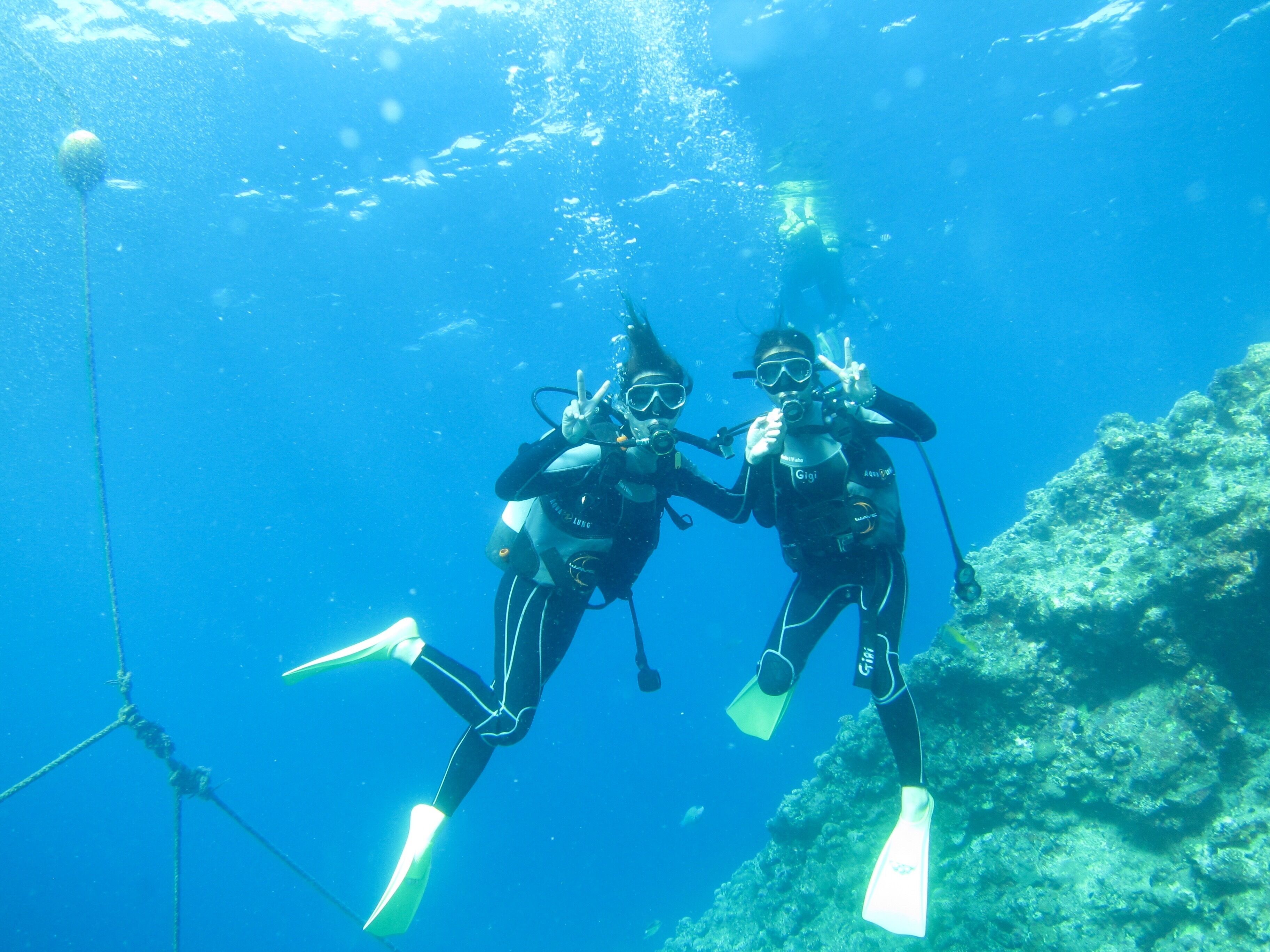 Dos buzos bajo el agua con aletas y equipo de buceo, rodeados de agua azul cristalina y un paisaje rocoso sumergido.