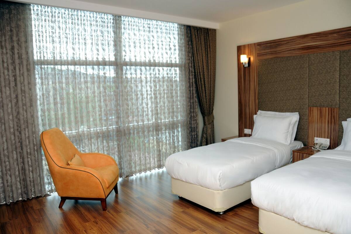 A hotel room with two beds, a wooden floor, a chair, and a large window with curtains.