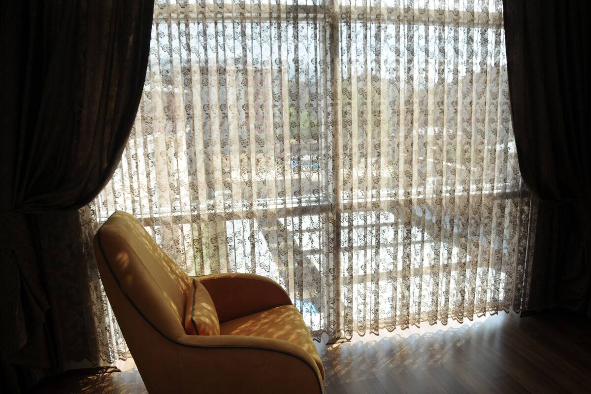 A room with a large window covered by sheer curtains, a wooden floor, and a single yellow armchair.