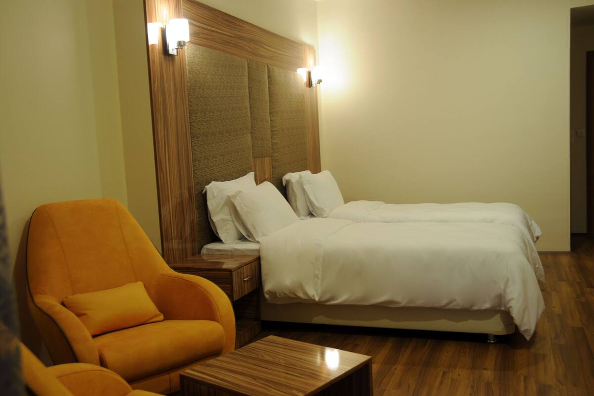 A hotel room with a bed, two pillows, a nightstand, a wooden headboard, a wooden floor, and an orange chair.