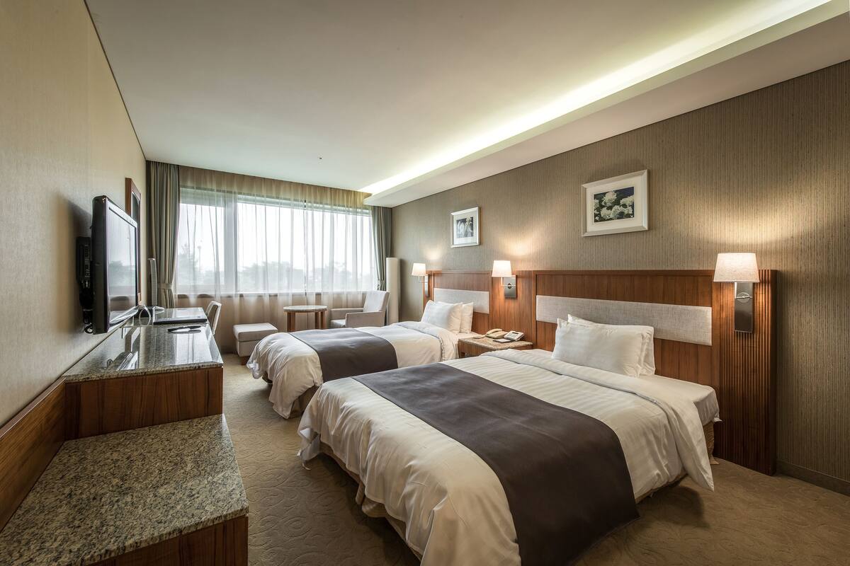 A hotel room with two beds, a desk, a television, and a window with curtains.