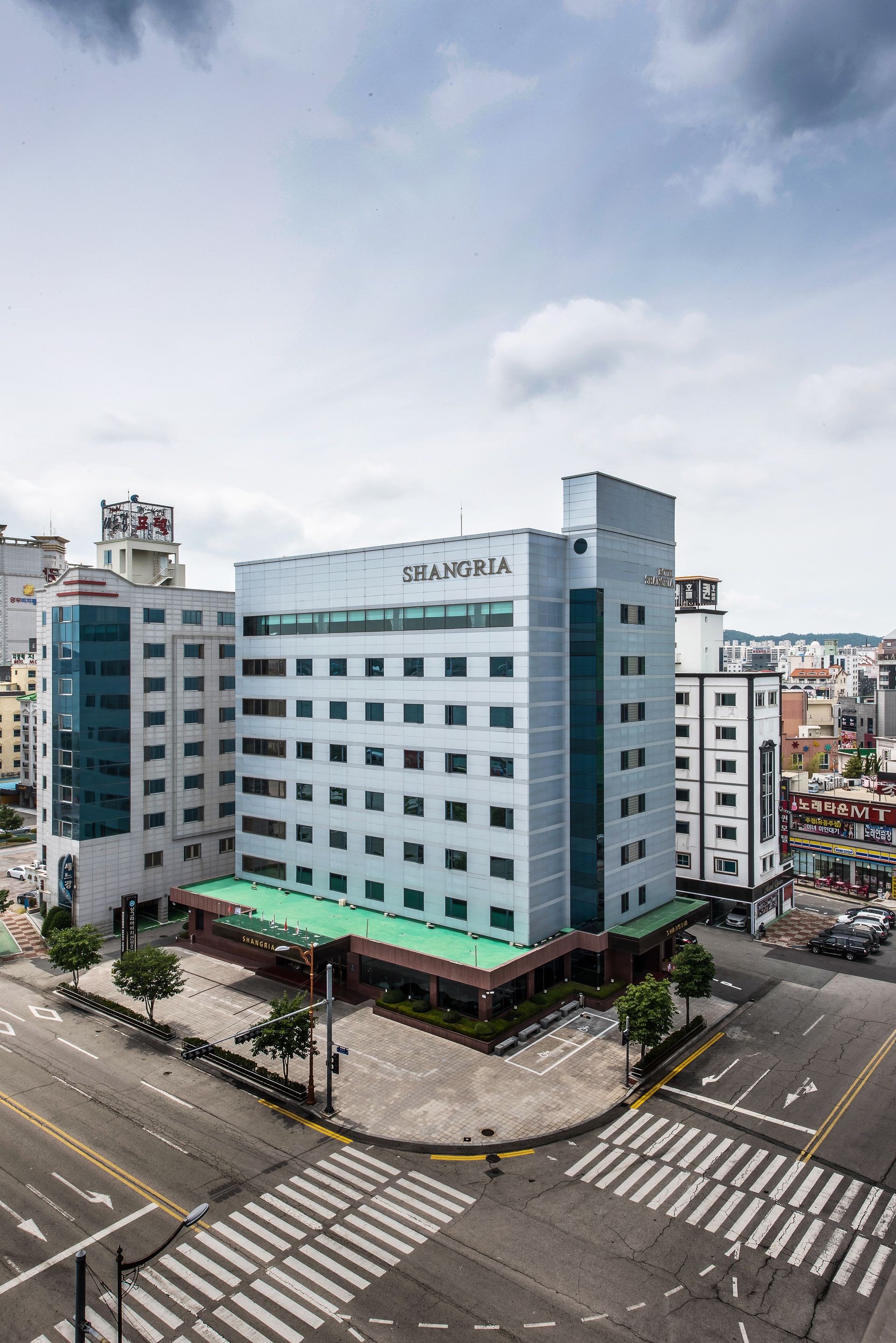 A multi-story building with a modern design, labeled "SHANGRIA." It features a green roof and is surrounded by a cityscape.