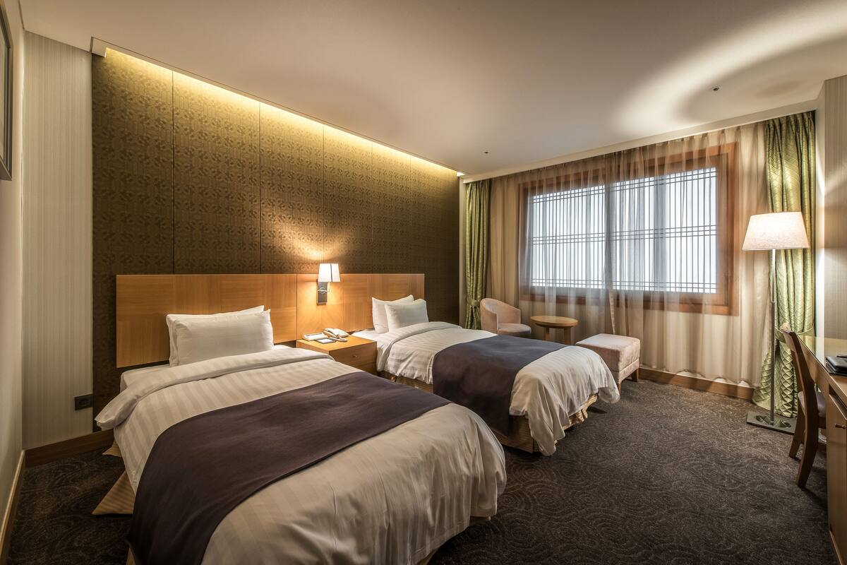 A hotel room with two beds, a desk, a chair, a lamp, and a window with curtains.