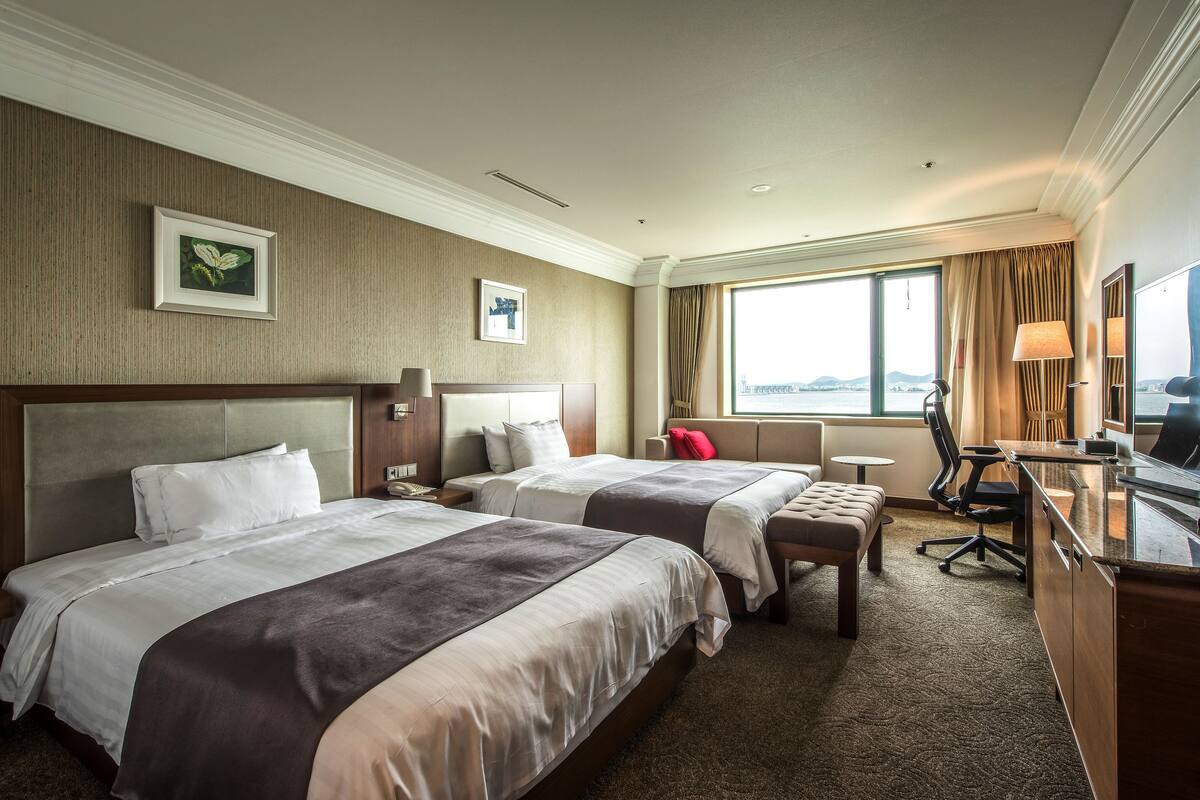 A hotel room with two beds, a desk, a chair, and a view of the city.
