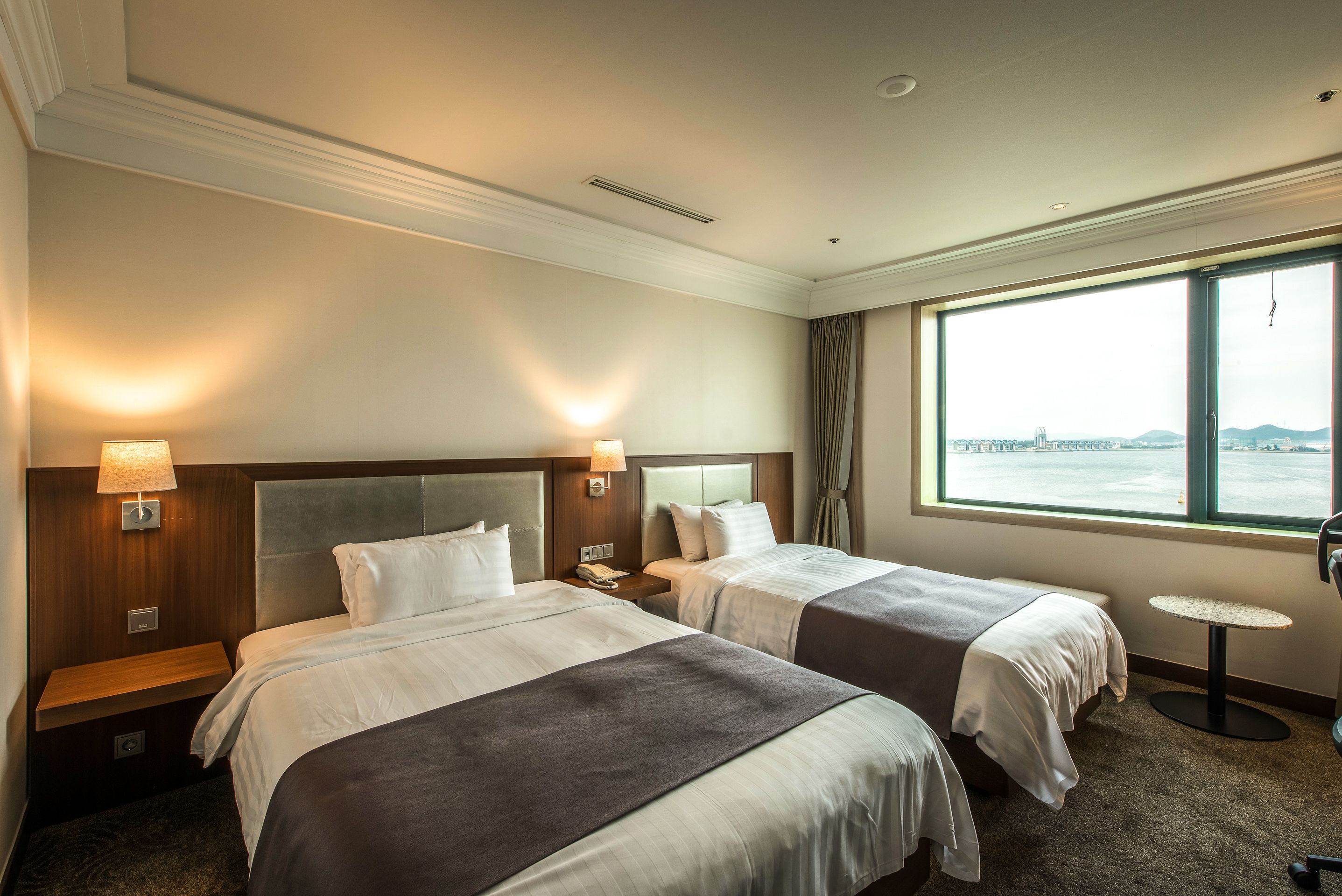 A hotel room with two beds, a small table, and a window with a city view.