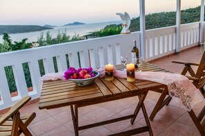 A wooden table with a bowl of fruit, candles, and a bottle on a balcony overlooking a body of water. - Anatoli Villa (Alonissos)