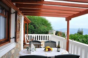 A covered outdoor dining area with a table set for two, a bottle of wine, and a fruit plate. - Anatoli Villa (Alonissos)