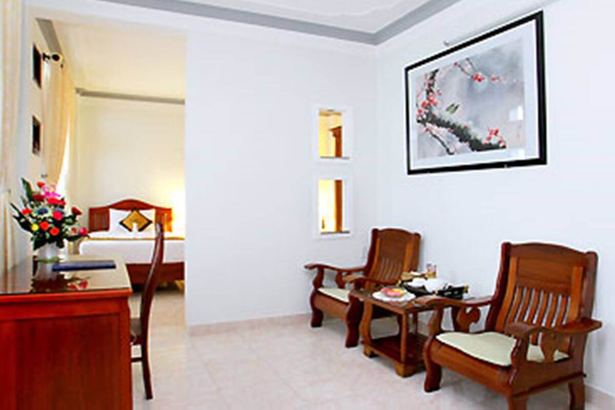 A hotel room with a bed, a desk with a vase of flowers, two wooden chairs, and a framed picture on the wall.