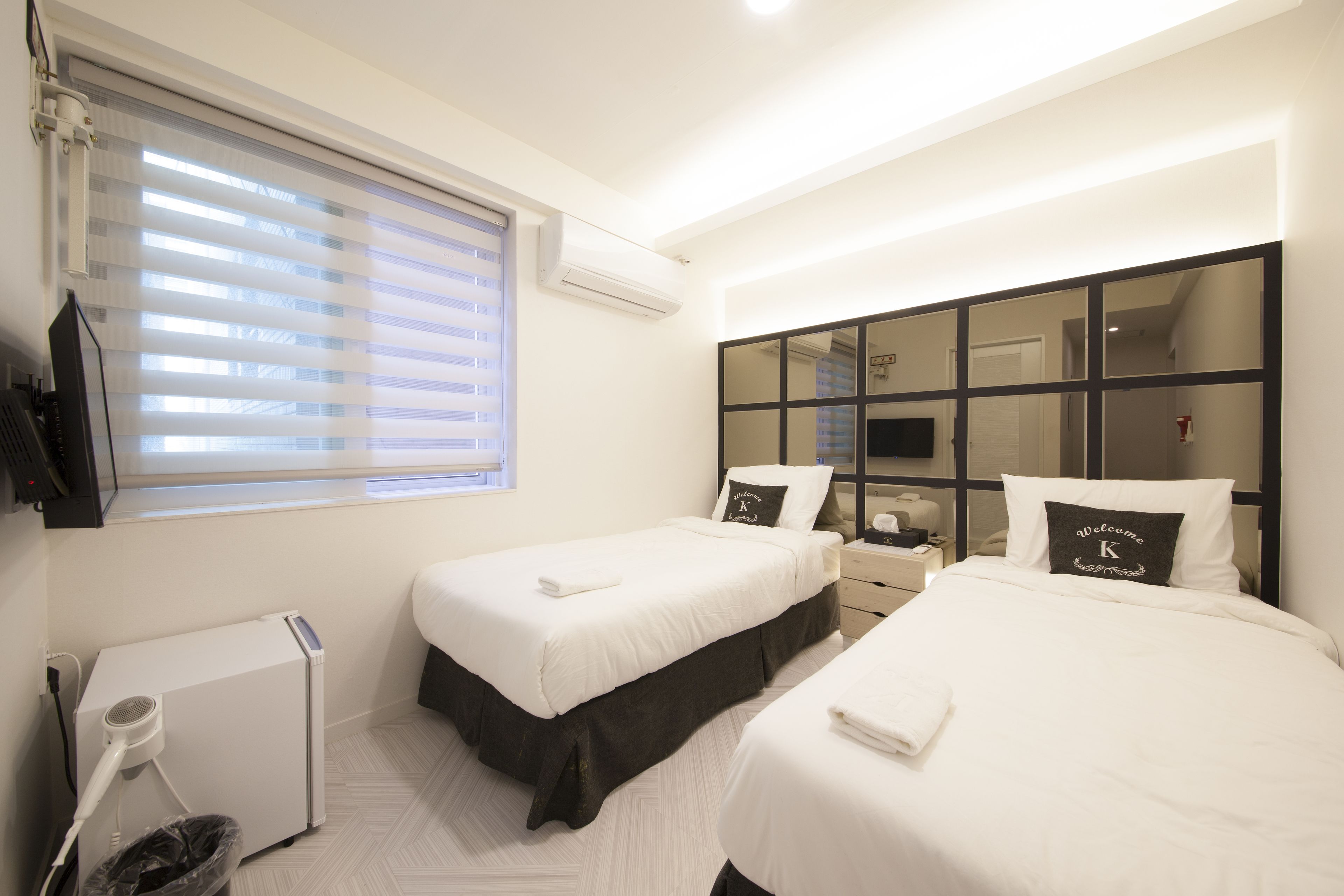 A hotel room with two beds, a large mirror, a hairdryer, and a window with blinds.