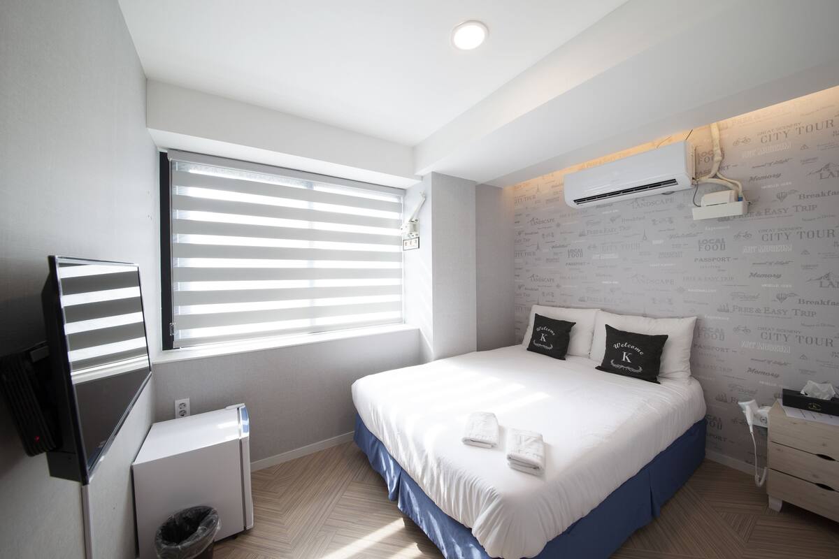 A modern hotel room with a large bed, a TV, a small desk, and a window with blinds.