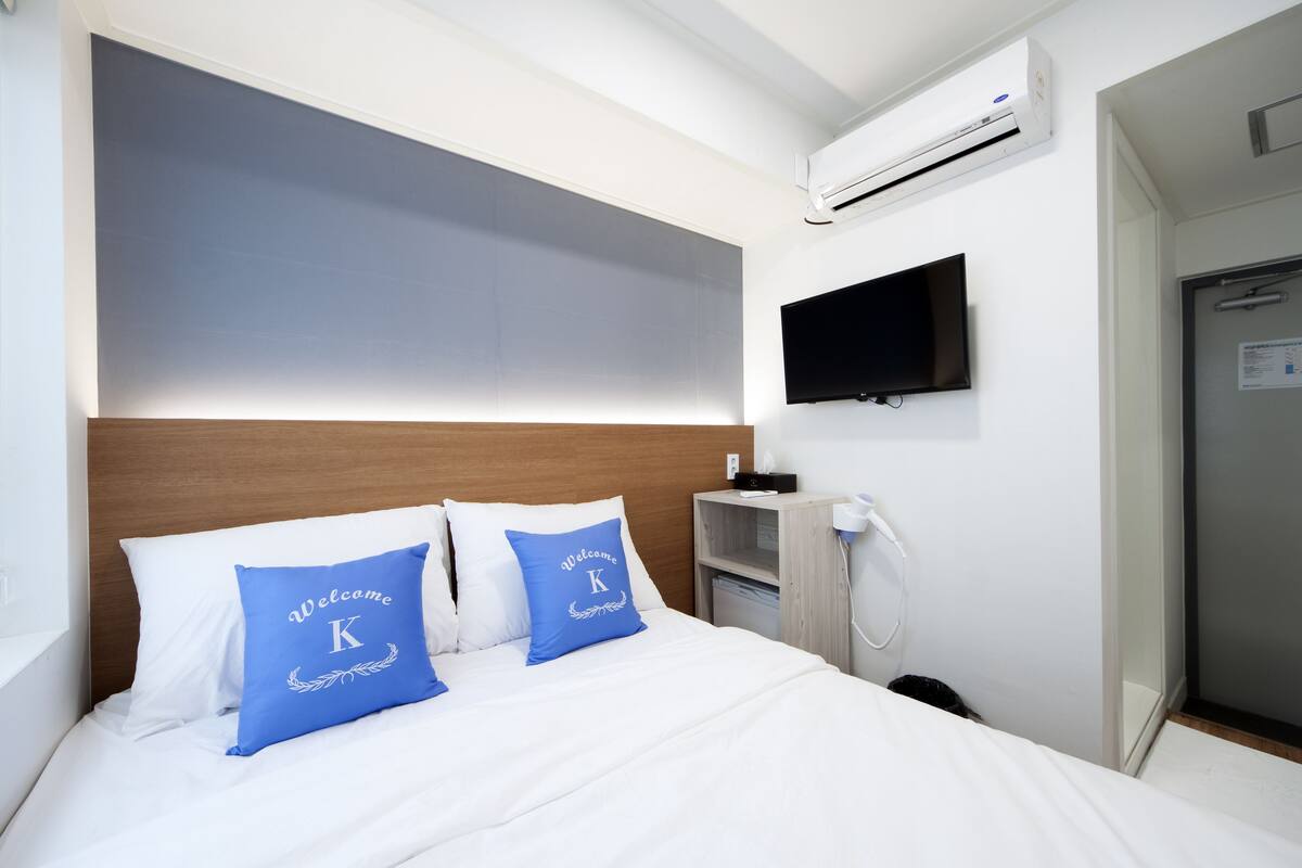 A hotel room with a bed, a TV mounted on the wall, and an air conditioning unit above the bed.