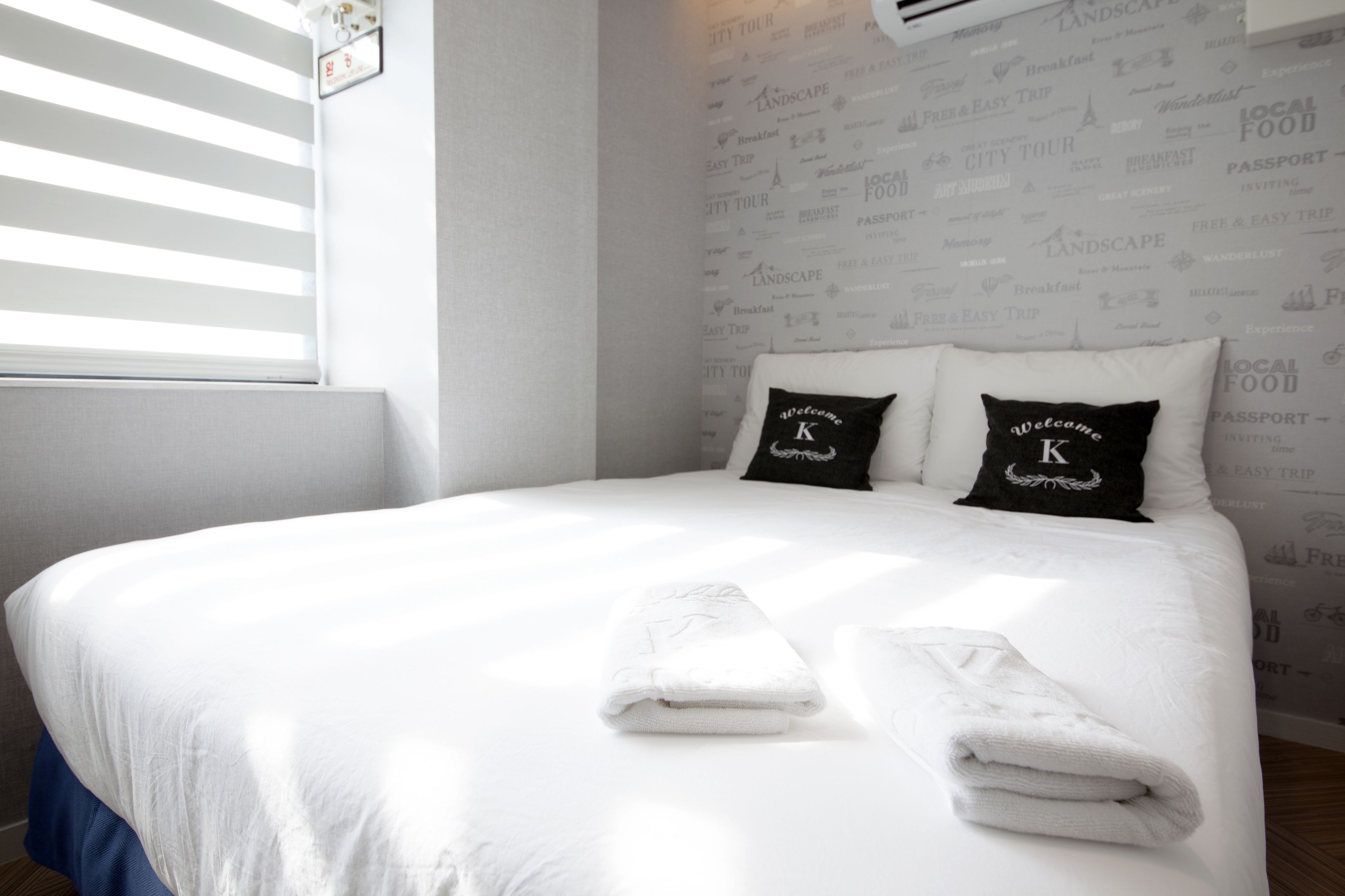 Cama bem arrumada com lençóis brancos e duas almofadas, uma com o emblema 'K', num quarto com papel de parede estampado e janela com persianas.