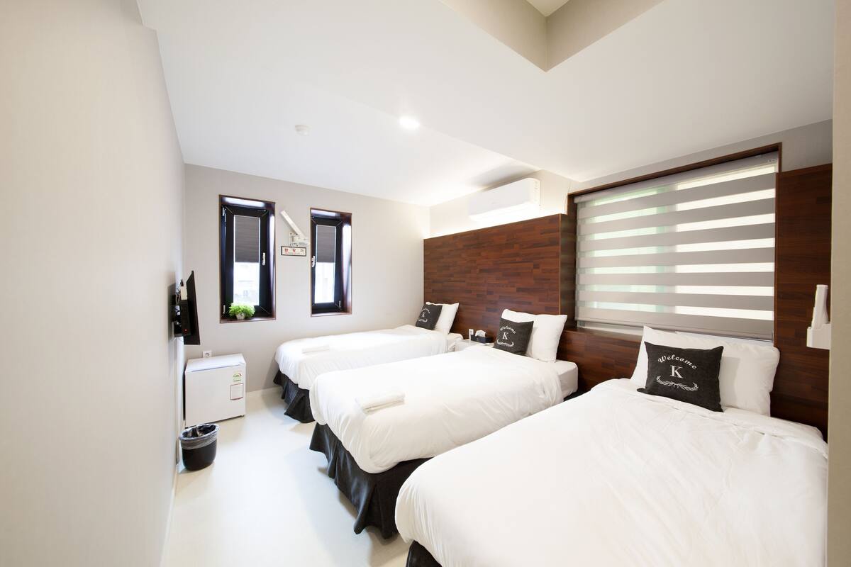 A hotel room with three beds, a small desk, and a window with blinds.