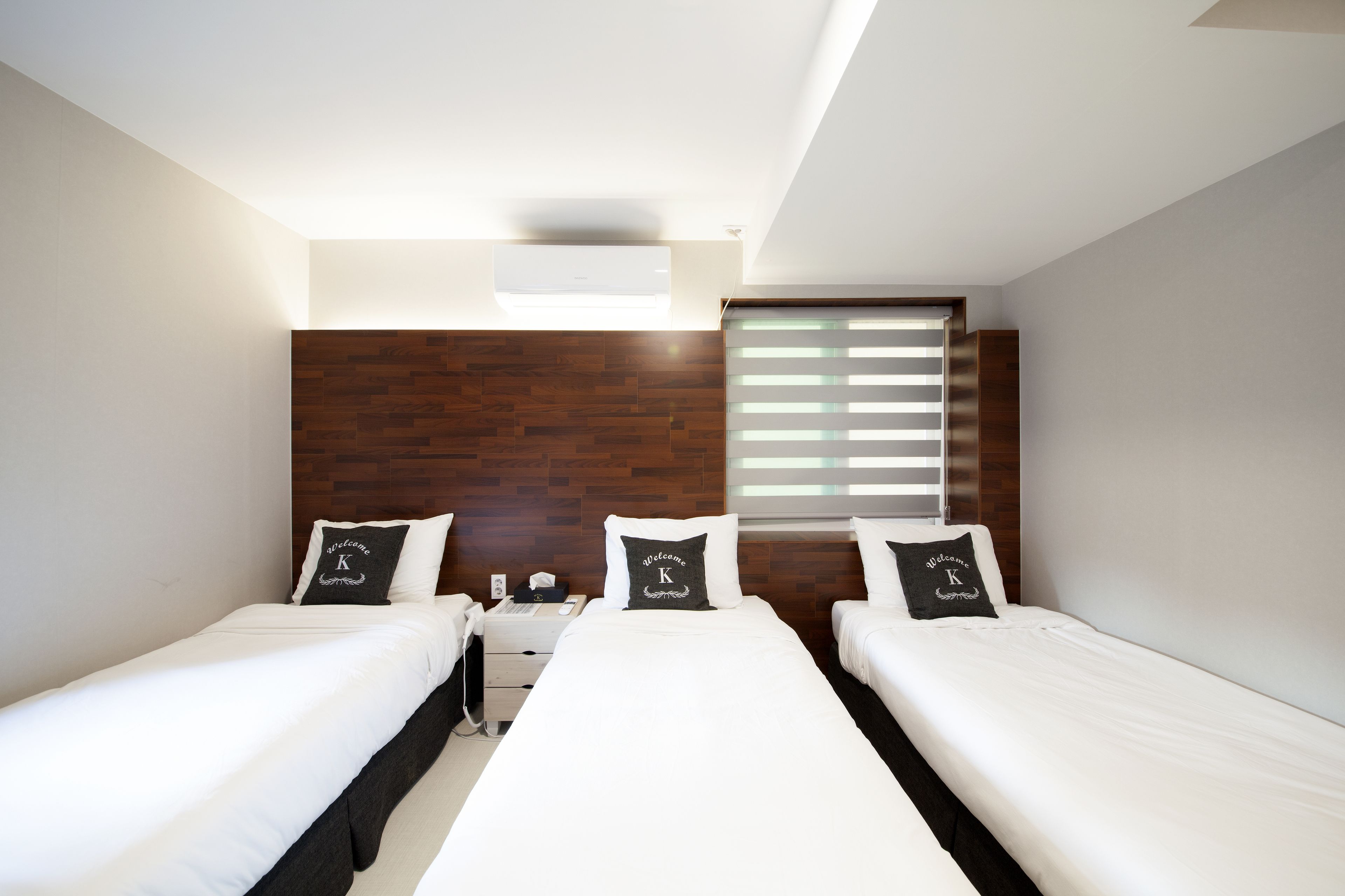 A modern hotel room with two single beds, a wooden headboard, a small bedside table, and a window with blinds.