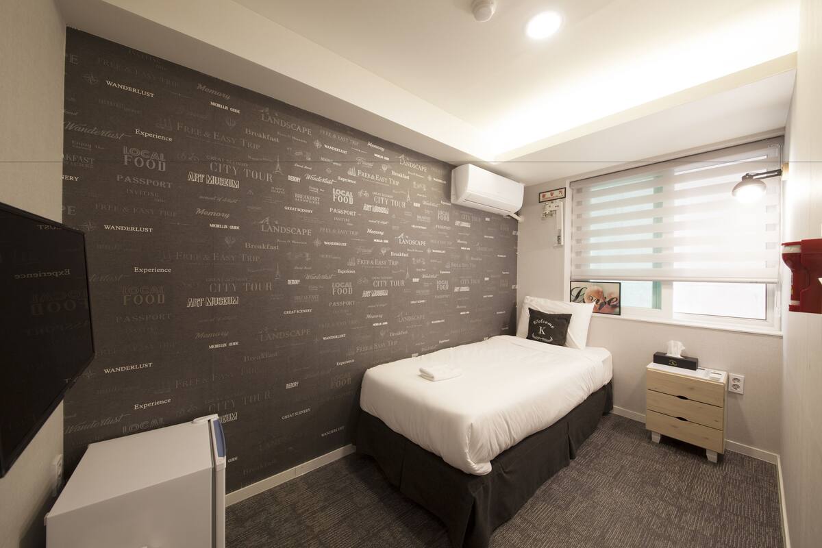 A hotel room with a bed, a bedside table, a wardrobe, a window with blinds, and a wall with various words and phrases.