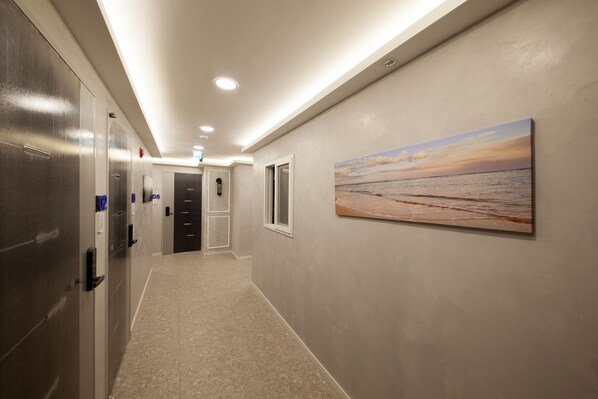 Pasillo de hotel con un cuadro de una playa al atardecer, un largo pasillo con puertas e iluminación empotrada en el techo.
