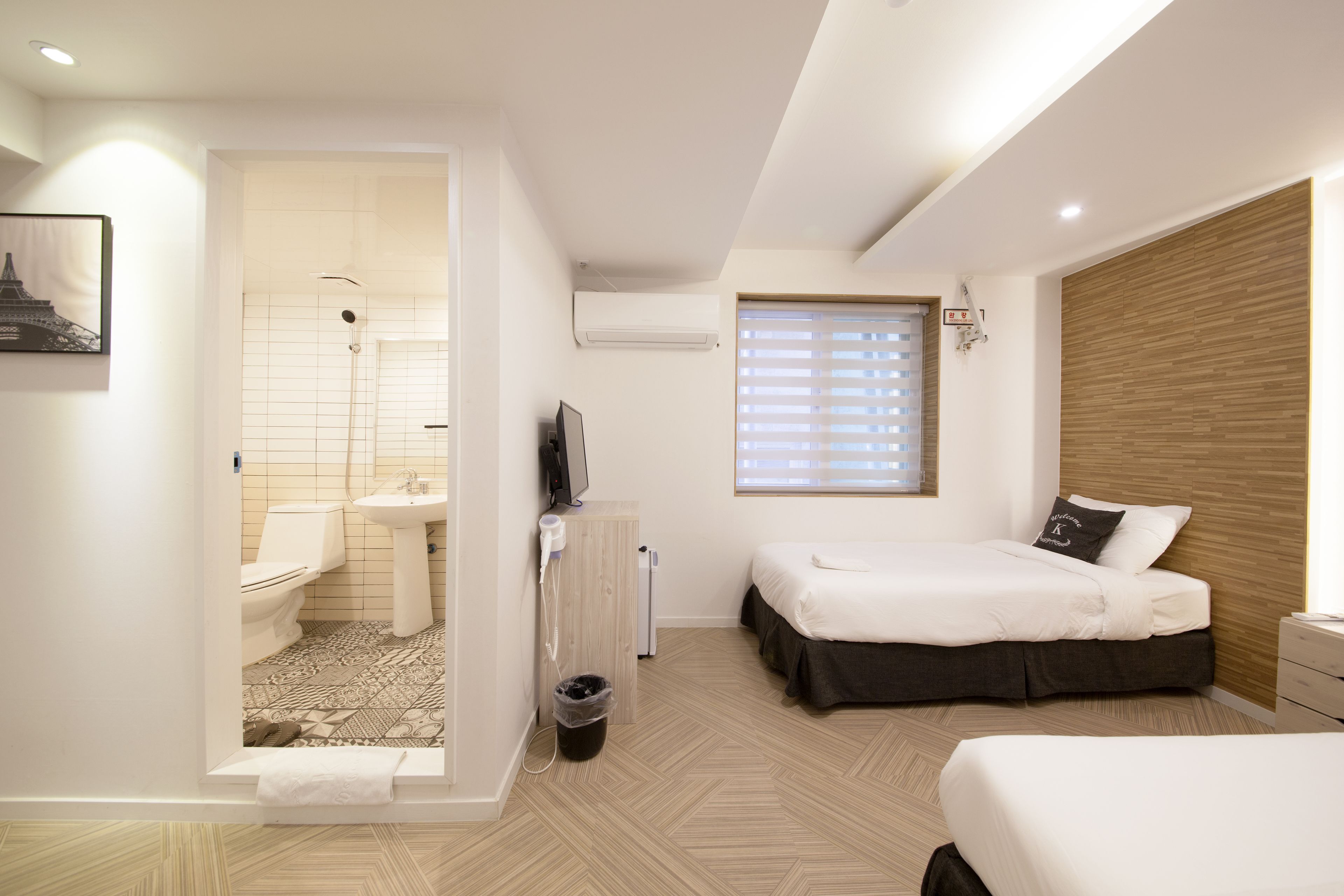 Ein modernes Hotelzimmer mit Bad, einem Bett mit Holz Kopfteil, einem Nachttisch und einem Fenster mit Jalousien.