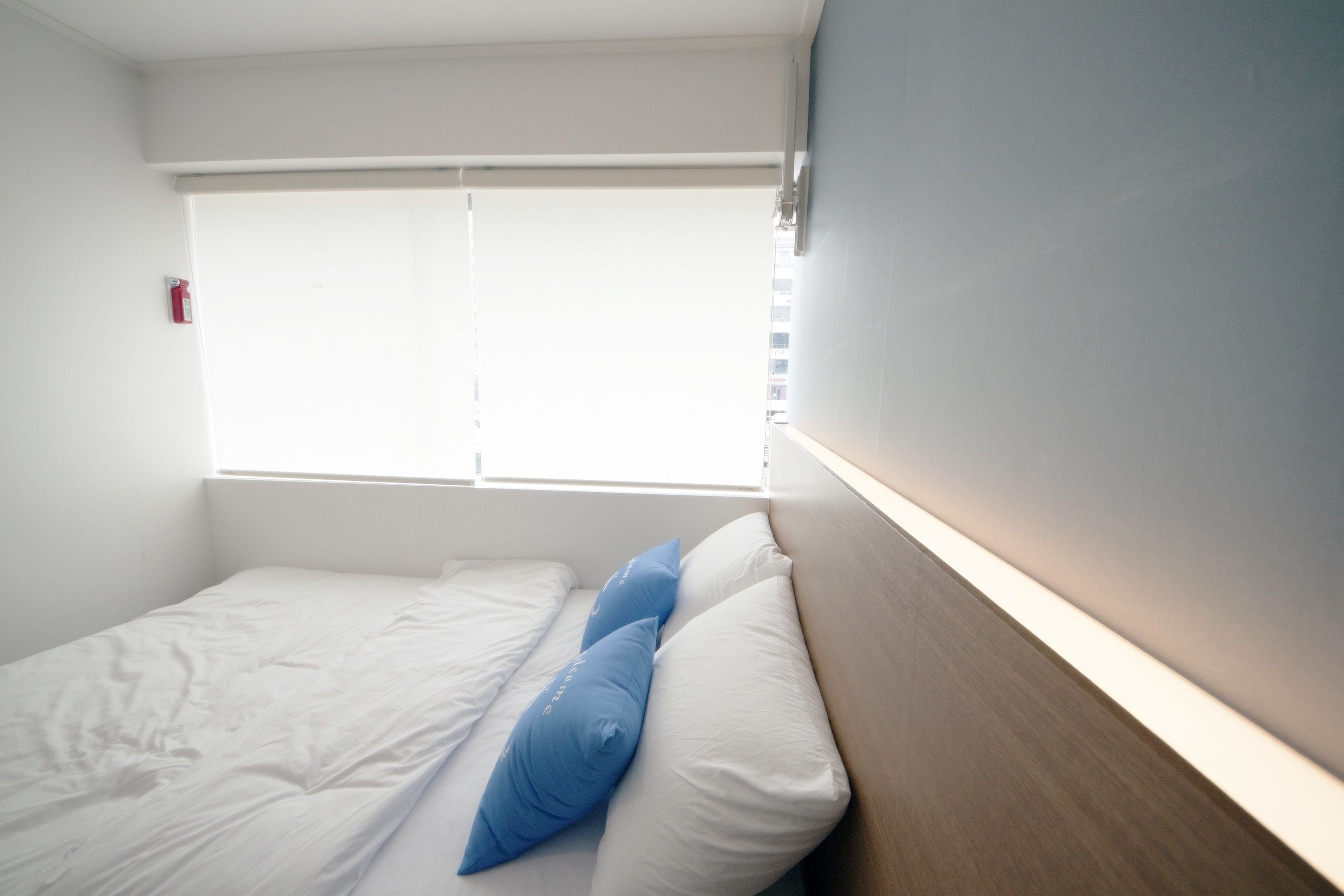 Una cama individual con almohadas blancas y azules, cabecera de madera y una ventana con persianas.