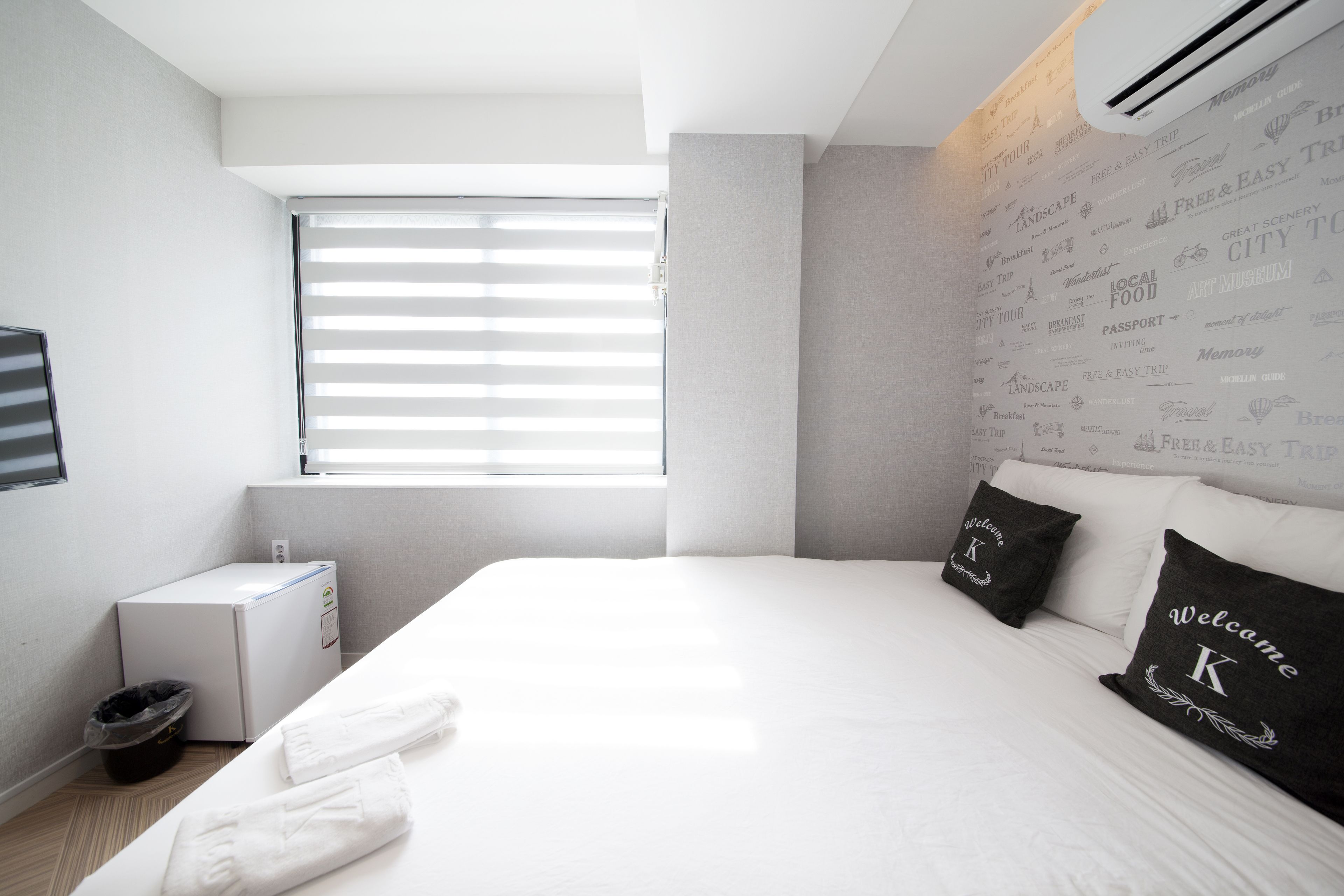 Une chambre d’hôtel moderne avec un grand lit, une petite table de chevet, une fenêtre avec des stores et un climatiseur fixé au mur.