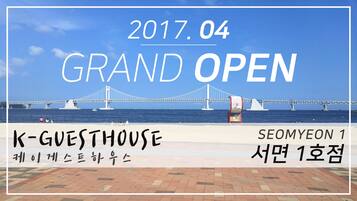 A grand opening event with a bridge in the background, a beach, and a sign for K-GUESTHOUSE.