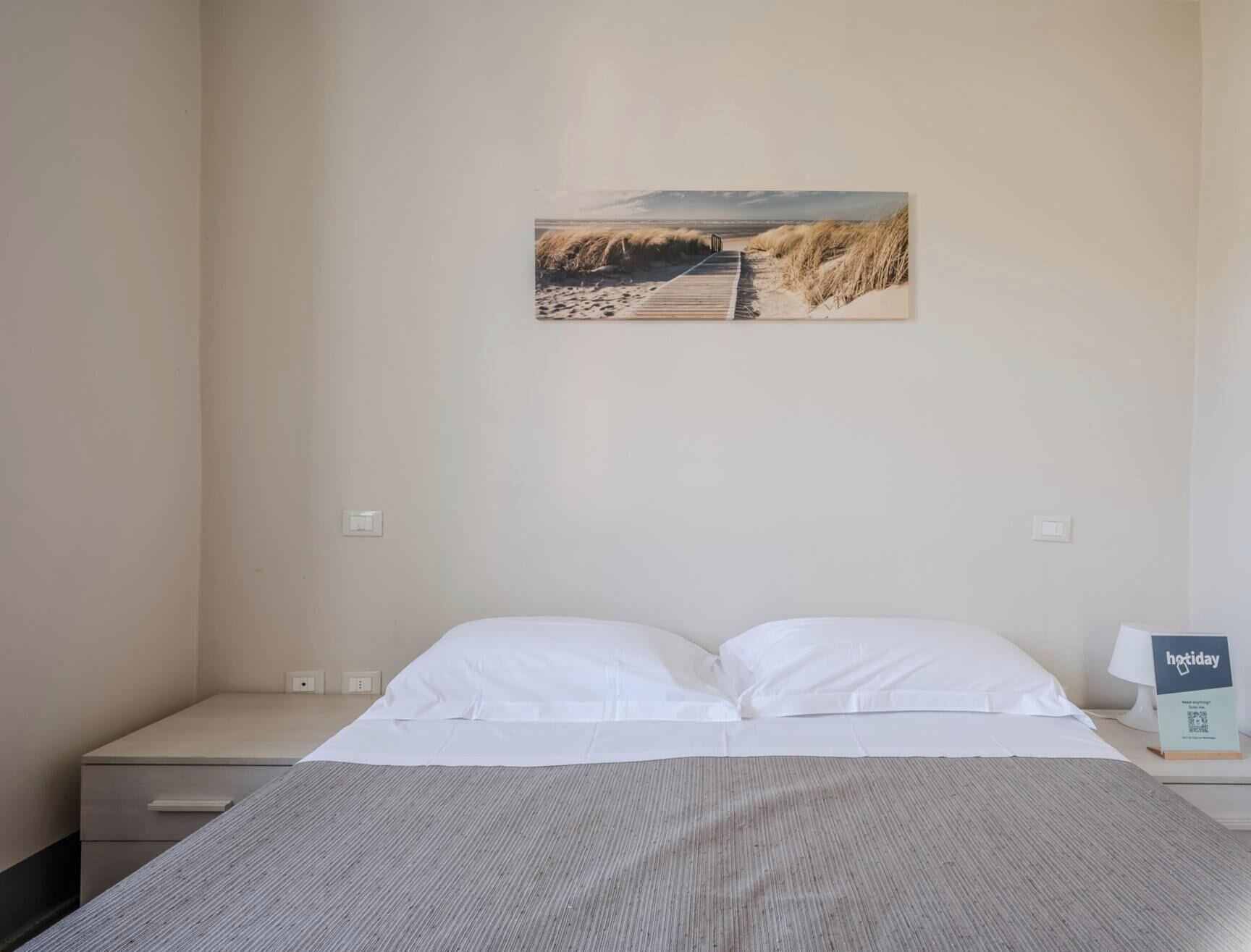 A bedroom with a bed, a nightstand, a lamp, and a wall-mounted picture of a beach.