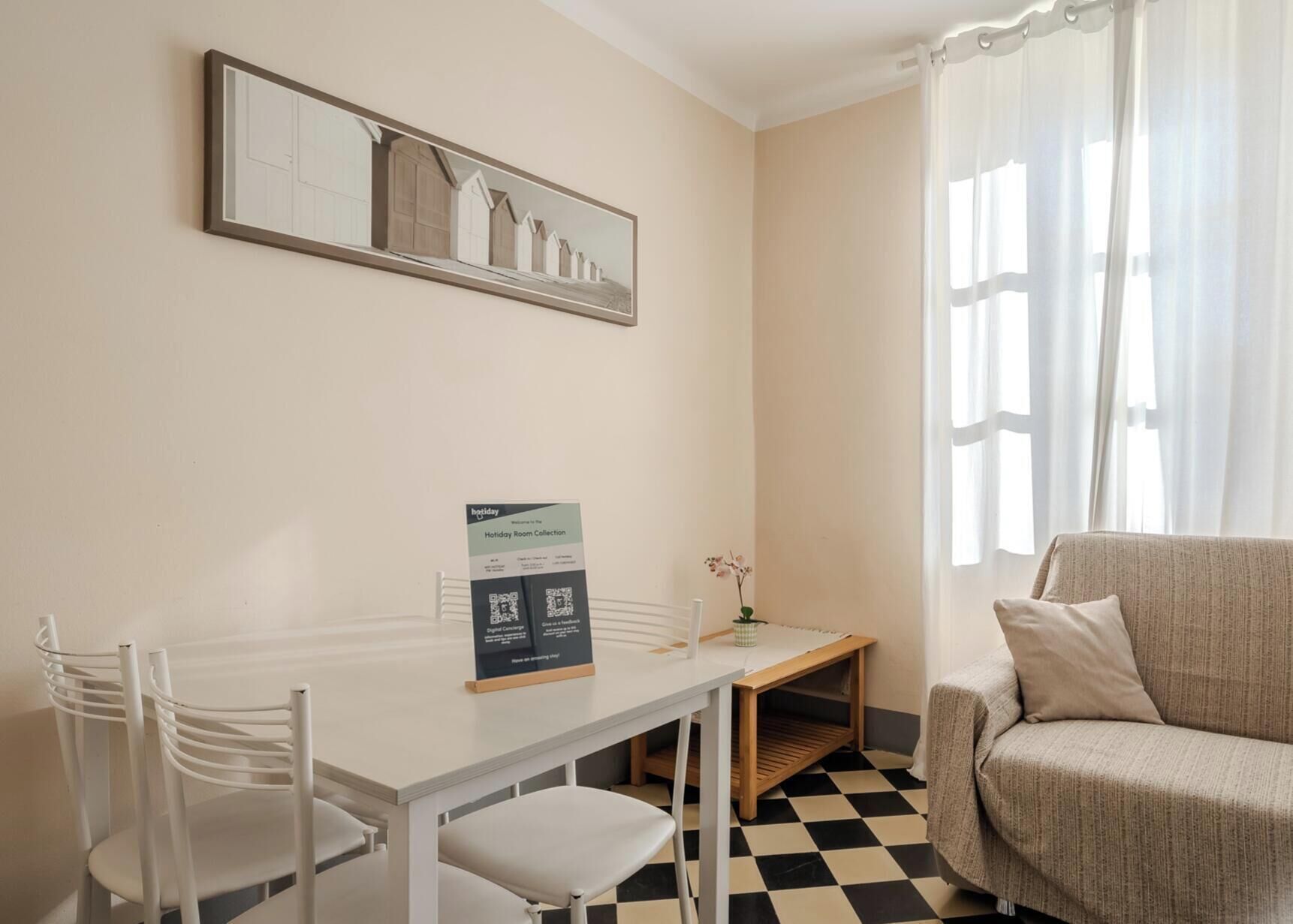 A hotel room with a checkered floor, a white desk with a sign, a chair, a sofa, and a painting on the wall.