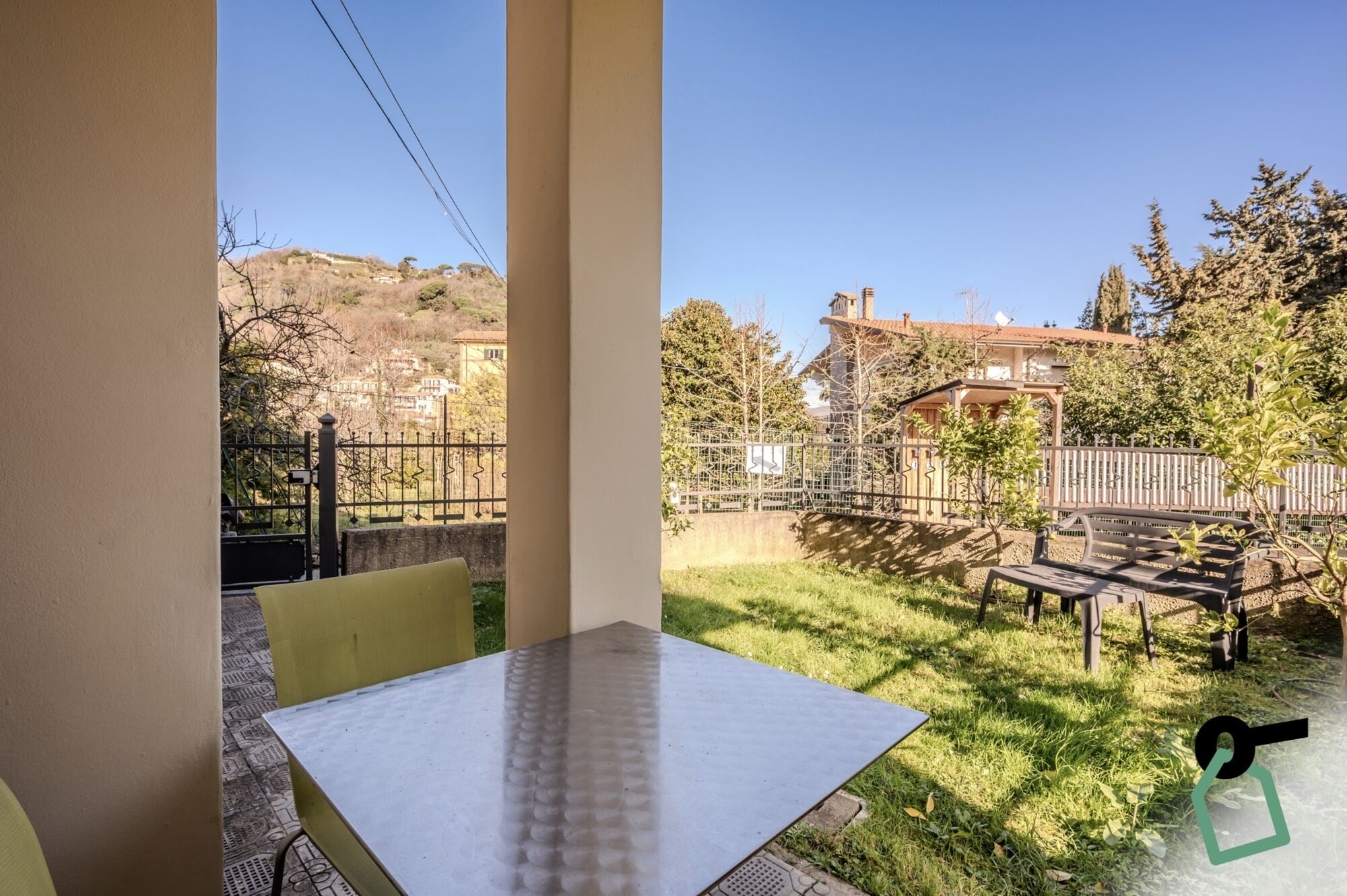 A balcony with a table and chairs, a view of a garden with a fence, and a building in the background.